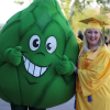A smiling graduate in a bright gold cap and gown stands outdoors beside a large green mascot shaped like a leafy plant with oversized cartoon eyes and a wide grin. The graduate has one arm around the mascot, and both appear to be giving a thumbs-up. In the background, other graduates and guests are gathered on a sunny campus lawn, creating a festive graduation atmosphere.