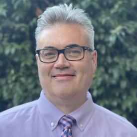 The subject with glasses, wearing a purple shirt and tie against a forest backdrop.